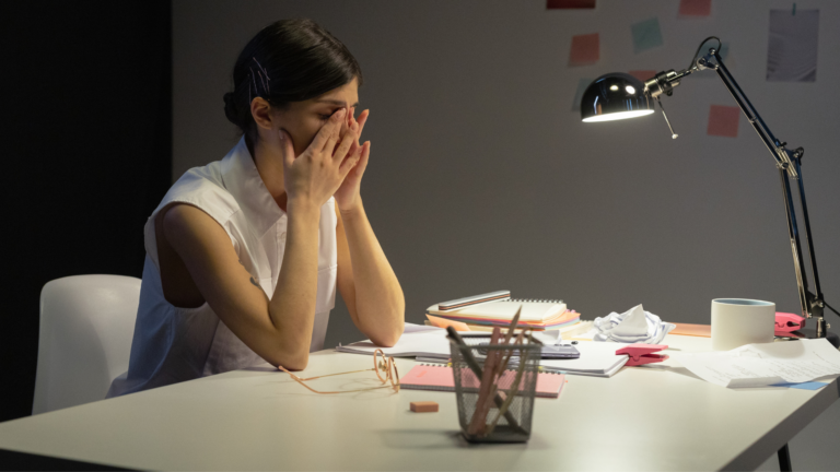 Professional woman sitting at her desk covering her face, reflecting feelings of workplace misalignment, stress, and emotional exhaustion while continuing to show up at work.