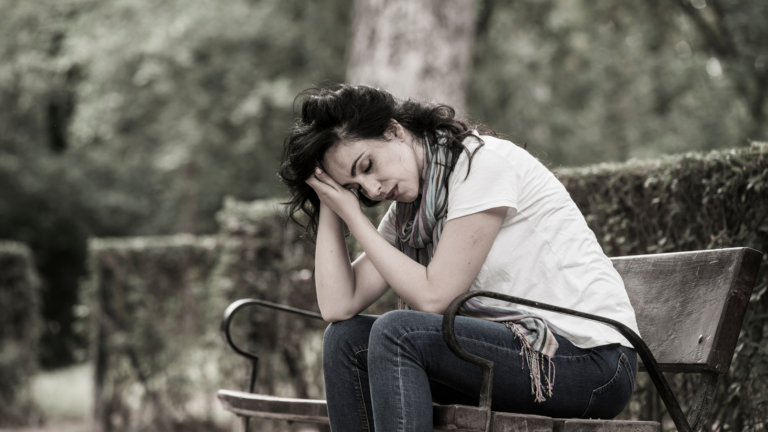 Woman sitting alone on a park bench with her head resting on her hands, appearing physically and emotionally exhausted.